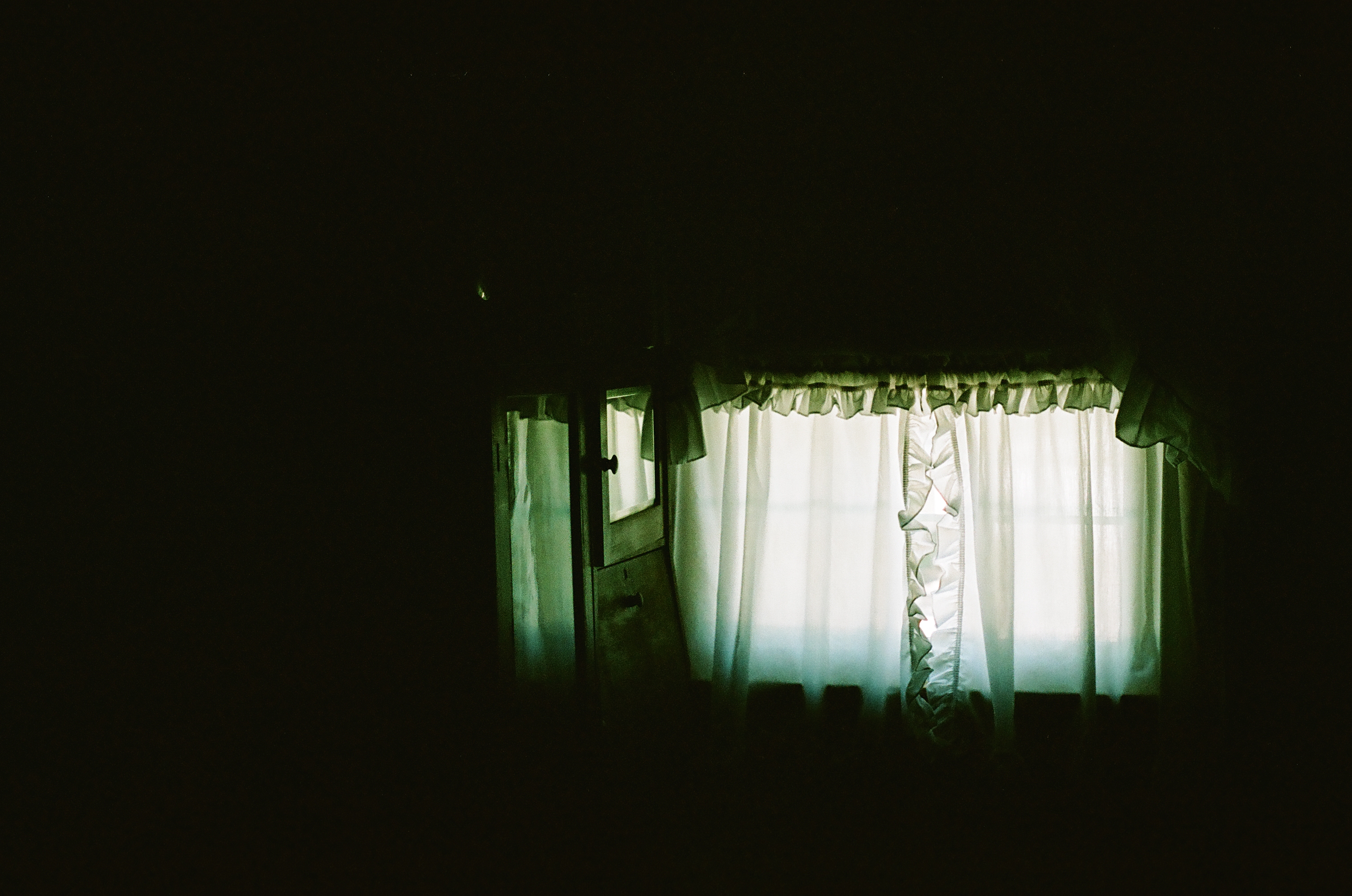 A dark interior room with sheer white curtains covering a bright window. The majority of the frame is in deep shadow.